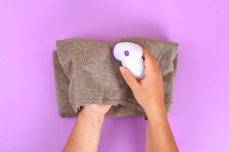 Canva - A Machine for Removing Lint in the Hand. Sweater with Bobble on a Purple Pastel Background.